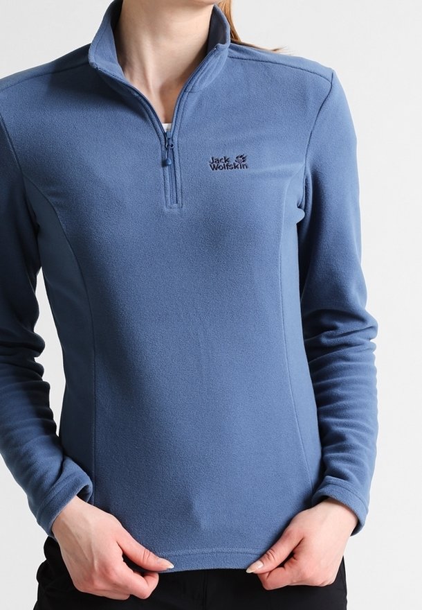 Femme portant un pull en polaire bleu Jack Wolfskin avec une fermeture éclair au quart, tenant l'ourlet du pull des deux mains.