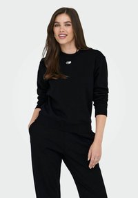 Γυναίκα με μακριά καστανά μαλλιά, φορώντας ένα μαύρο μπλουζάκι με μακριά μανίκια και παντελόνι, χαμογελαστή με το ένα χέρι στην τσέπη, μπροστά από μια απλή φωτεινή φόντο.