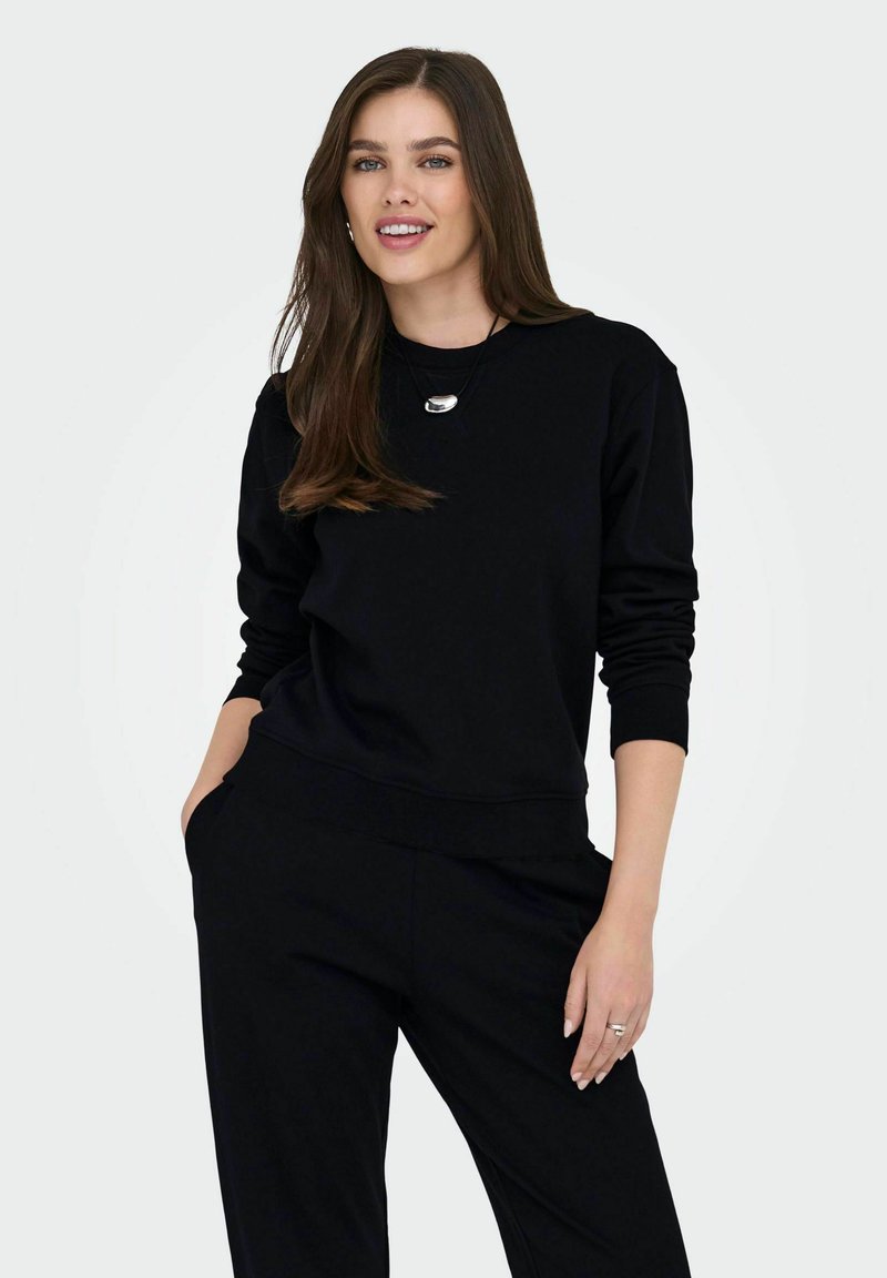 Γυναίκα με μακριά καστανά μαλλιά, φορώντας ένα μαύρο μπλουζάκι με μακριά μανίκια και παντελόνι, χαμογελαστή με το ένα χέρι στην τσέπη, μπροστά από μια απλή φωτεινή φόντο.