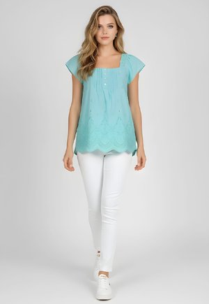 Mujer con cabello suelto y ondulado que lleva una blusa sin mangas turquesa con patrón de encaje y pantalones blancos ajustados caminando hacia adelante sobre un fondo liso.