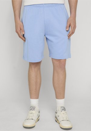 Mand iført lyseblå elastiske shorts i taljen, hvide crew-sokker og hvide New Balance sneakers, stående mod en ensfarvet grå baggrund.