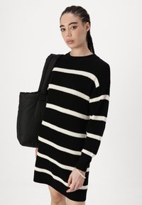Vestido tricotado de riscas pretas e brancas com mangas longas, gola redonda e textura canelada. Uma bolsa tote preta está pendurada no ombro.