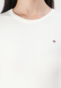 White ribbed top with a round neckline and short sleeves, featuring a small embroidered logo in red, white, and blue on the left chest.