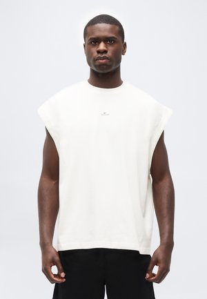 Man draagt een mouwloos wit Nike-top en zwarte broek, staand met het gezicht naar voren tegen een effen lichte achtergrond.