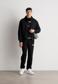 Czarna kurtka bomberka noszona na czarnej bluzie z kapturem, w zestawieniu z czarnymi spodniami dresowymi z białym logo. Białe sneakersy i szara torba na ramię.