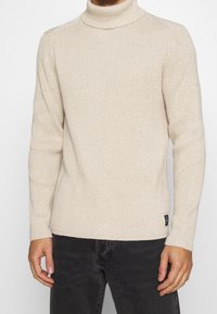 Maglione a collo alto beige a coste con maniche lunghe e design aderente. Presenta un piccolo tag con logo nero in basso a sinistra.