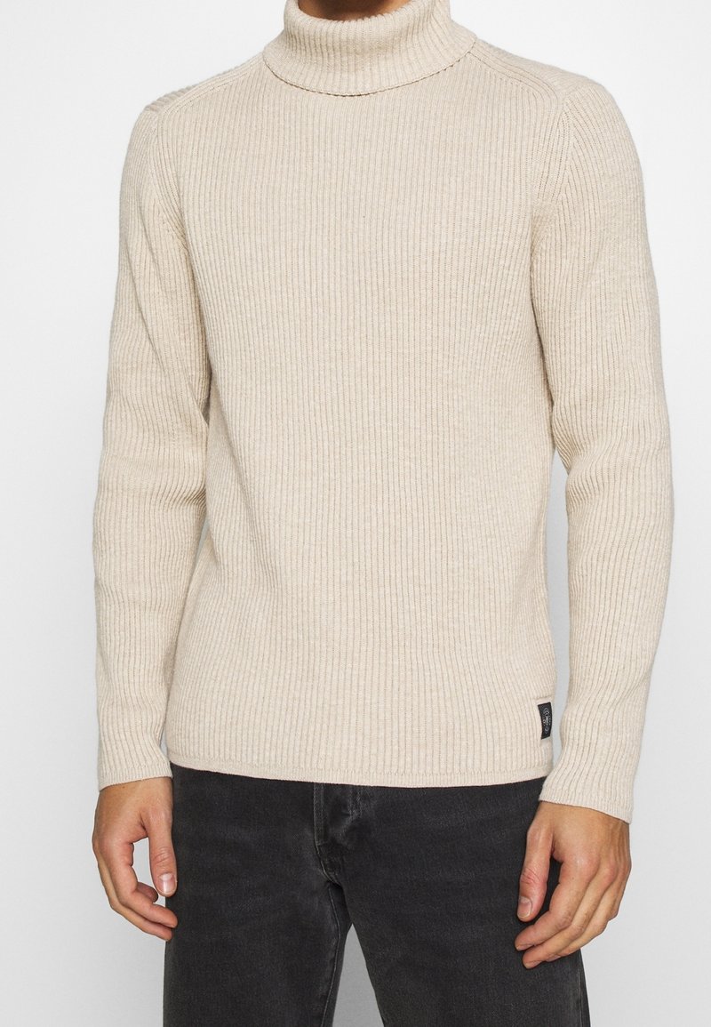Maglione a collo alto beige a coste con maniche lunghe e design aderente. Presenta un piccolo tag con logo nero in basso a sinistra.
