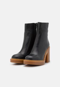 Botas de cuero negro hasta el tobillo con un tacón alto y grueso y un acabado suave, que cuentan con una cremallera lateral y una suela beige en contraste.
