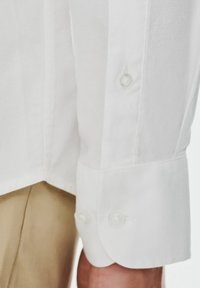 Biała tkanina koszuli z teksturowanym wykończeniem, z zaokrąglonymi mankietami z dwoma jasnymi guzikami. Zbliżenie pokazuje dół koszuli i sąsiednie beżowe spodnie.