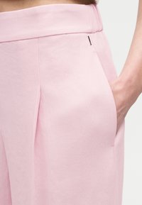 Κοντινό πλάνο ατόμου που φοράει ανοιχτό ροζ παντελόνι με λεπτομέρεια πτυχώματος, το χέρι ακουμπάει στην πλαϊνή τσέπη, με φόντο ένα απλό φόντο.