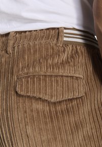 Brune kordfløjspants med lodrette ribber, med en baglomme og en stribet elastisk talje i brun og hvid.