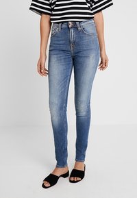 Modré džíny s vysokým pasem a slim fit, s efektivním nebo poškozeným vzhledem na kolenou. Kombinováno s černými nazouvacími sandály.