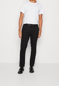 Schwarze, schmal geschnittene Hose mit flacher Vorderseite, kombiniert mit einem weißen T-Shirt mit Rundhalsausschnitt und schwarzen Schuhen. Verfügt über einen schwarzen Gürtel mit einer runden Schnalle.