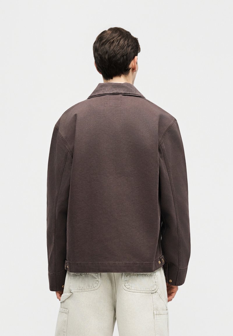 Hombre de espaldas que lleva una chaqueta marrón oscuro y pantalones cargo claros contra un fondo liso y claro.