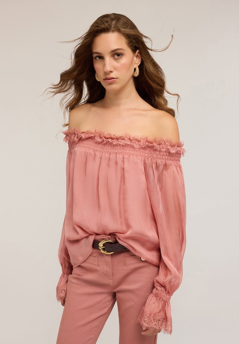 Blusa rosa trasparente off-the-shoulder con bordo arricciato e polsini arricciati, abbinata a pantaloni rosa e una cintura marrone con fibbia dorata.