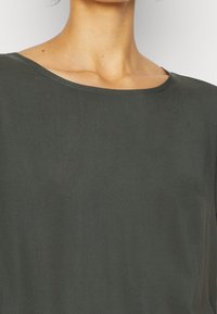 Top verde scuro con scollatura rotonda, maniche corte e un tessuto liscio e leggermente strutturato. Design semplice e minimalista.