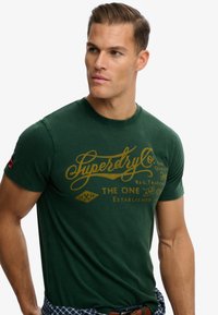 Grøn bomulds T-shirt med et guldtryk af logo og tekst på fronten, korte ærmer og rund halsudskæring.