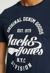Camiseta azul marino con texto blanco que dice "Original Denim Goods Est. 1975 Jack & Jones N.Y.C Division" en el pecho.