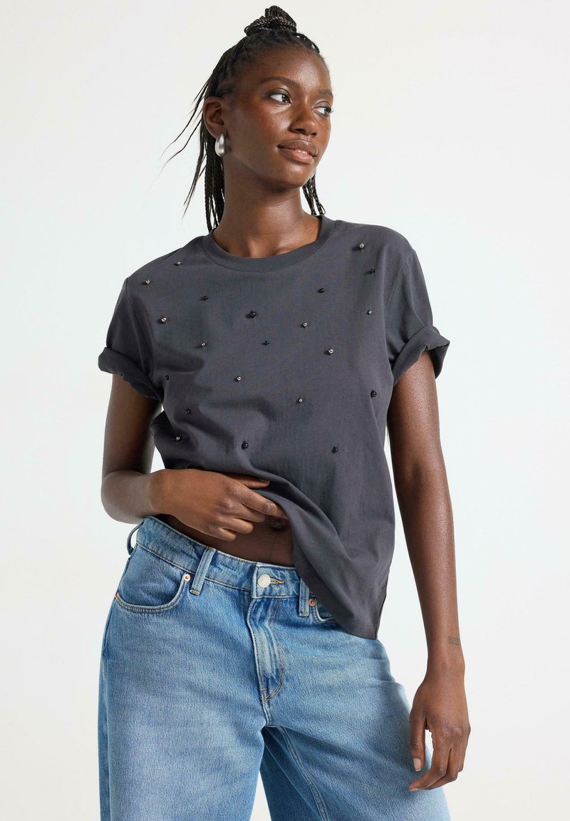 Mørkegrå kortærmet t-shirt med opsmøgte ærmer, prydet med små juvelliknende udsmykninger i varierende størrelser. Matchet med lys blå jeans.