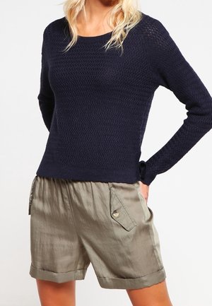 Vrouw draagt een marineblauwe, gestructureerde trui met lange mouwen en lichte olijfgroene, losse korte broek met voorzakken met knopen.