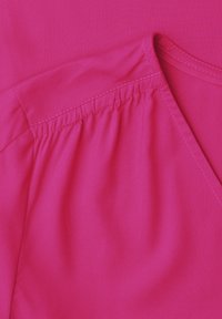 Vêtement rose vif avec des coutures froncées le long de la couture d'épaule et un col arrondi avec surpiqûres visibles.