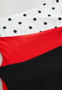 Koszulka wykonana z białej tkaniny w czarne kropki, z czerwonymi i czarnymi sekcjami. Gładka tekstura, warstwowy design z wyraźnym kontrastem kolorystycznym.