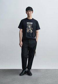 Μαύρο βαμβακερό μπλουζάκι με γραφικό αρκουδάκι και κείμενο «Moschino», σετ με μαύρο παντελόνι που έχει πλαϊνό φερμουάρ και μαύρα αθλητικά παπούτσια.