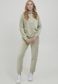 Ljusgrön velour träningsdräkt med en halv dragkedja på pullovern och matchande kilformade byxor. Har elastiska muddar och en mjuk textur. Vita sneakers.