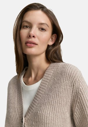 Frau mit langen braunen Haaren, die eine beigen Strickjacke über einem weißen Shirt trägt und leicht in Richtung Kamera blickt, vor einem weißen Hintergrund.