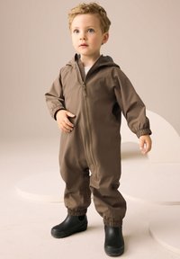 Combinaison imperméable marron avec fermeture éclair à l'avant, poignets élastiques et capuche ; portée avec des bottes en caoutchouc noires sur un fond clair.