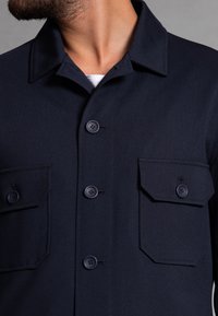 Navy blue work jacket made of textured fabric, featuring a pointed collar, button front, and two chest pockets with flap closures.