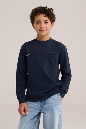 Un enfant aux cheveux bouclés porte un sweat-shirt bleu marine à col rond et un jean bleu clair sur un fond beige uni.
