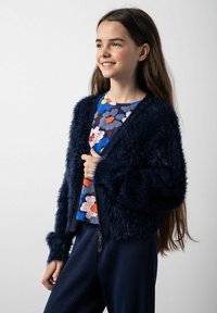 Granatowy, puszysty kardigan z szerokimi rękawami, noszony na kolorowej bluzce w kwiatowy wzór. Materiał wydaje się być miękki i teksturowany, dopełniony prążkowanymi spodniami.