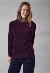 Felpa in corduroy viola con maniche lunghe, texture a coste e vestibilità rilassata, abbinata a jeans scuri. Presenta un sottile accento sull'orlo.