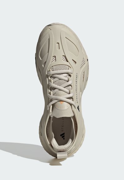 Zapatilla deportiva beige con parte superior de malla texturizada, que presenta un revestimiento de plástico moldeado, punta redonda y acentos de logo en contraste.