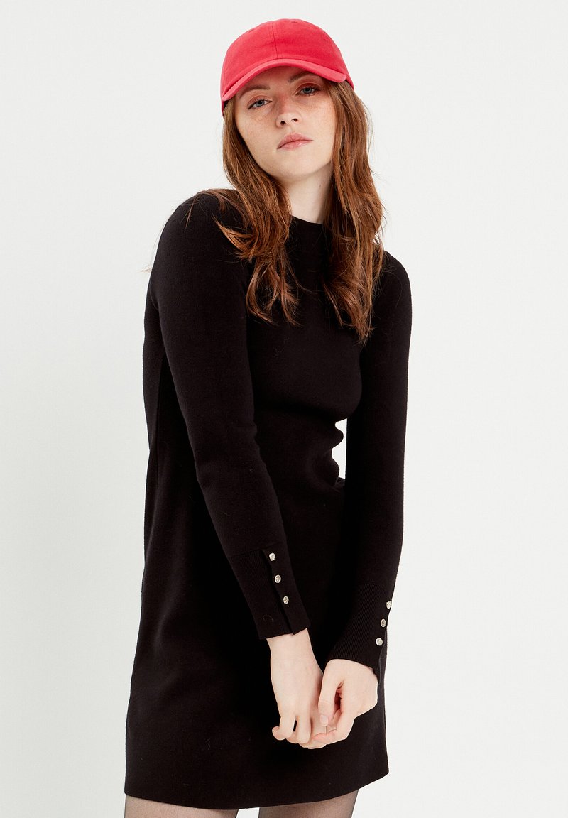 Robe en tricot noire à manches longues, avec des boutons décoratifs aux poignets. Associée à une casquette de base-ball rouge. Texture lisse et design ajusté.