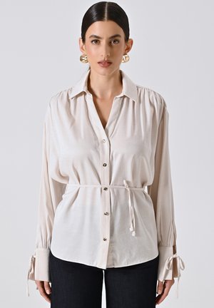Camicia button-up beige chiaro con maniche lunghe, vita stretta e polsini legati. Tessuto liscio con una leggera lucentezza e dettagli con bottoni frontali.