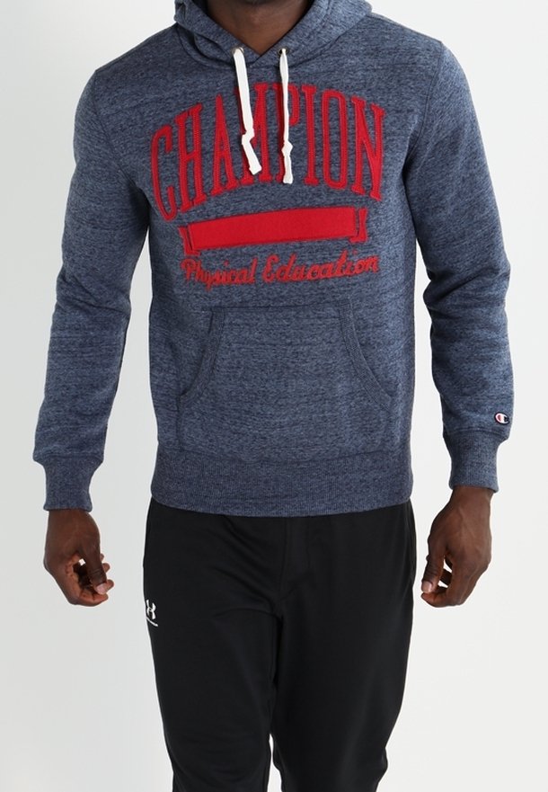 Homme portant un hoodie Champion bleu avec le texte rouge "CHAMPION Physical Education" et un pantalon noir Under Armour, debout devant un fond blanc.