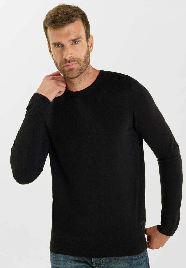 Uomo con capelli corti castani e barba, che indossa un maglione nero a maniche lunghe, mentre posa con una mano vicino al collo e l'altra sui fianchi.