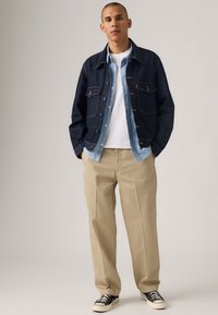 Dark denim jacket over a light blue shirt, paired with beige trousers and black sneakers. Features two chest pockets and contrasting buttons.