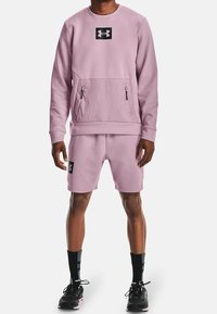 Felpa sportiva rosa con tasca frontale e zip nere, abbinata a pantaloni corti rosa con toppa nera. Scarpe da ginnastica nere.