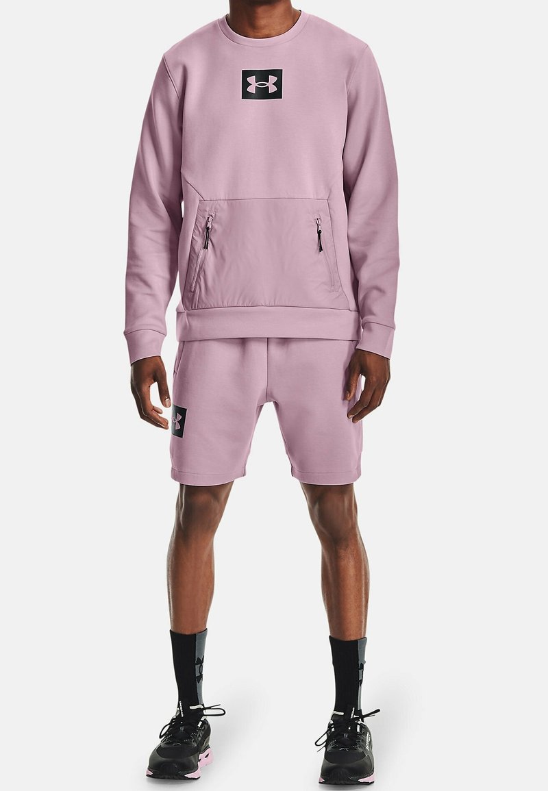 Felpa sportiva rosa con tasca frontale e zip nere, abbinata a pantaloni corti rosa con toppa nera. Scarpe da ginnastica nere.