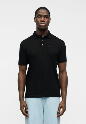 Muž oblečený v čiernej polokošeli s krátkym rukávom a svetlomodrých džínsoch, stojaci pred jednoduchým bielym pozadím.