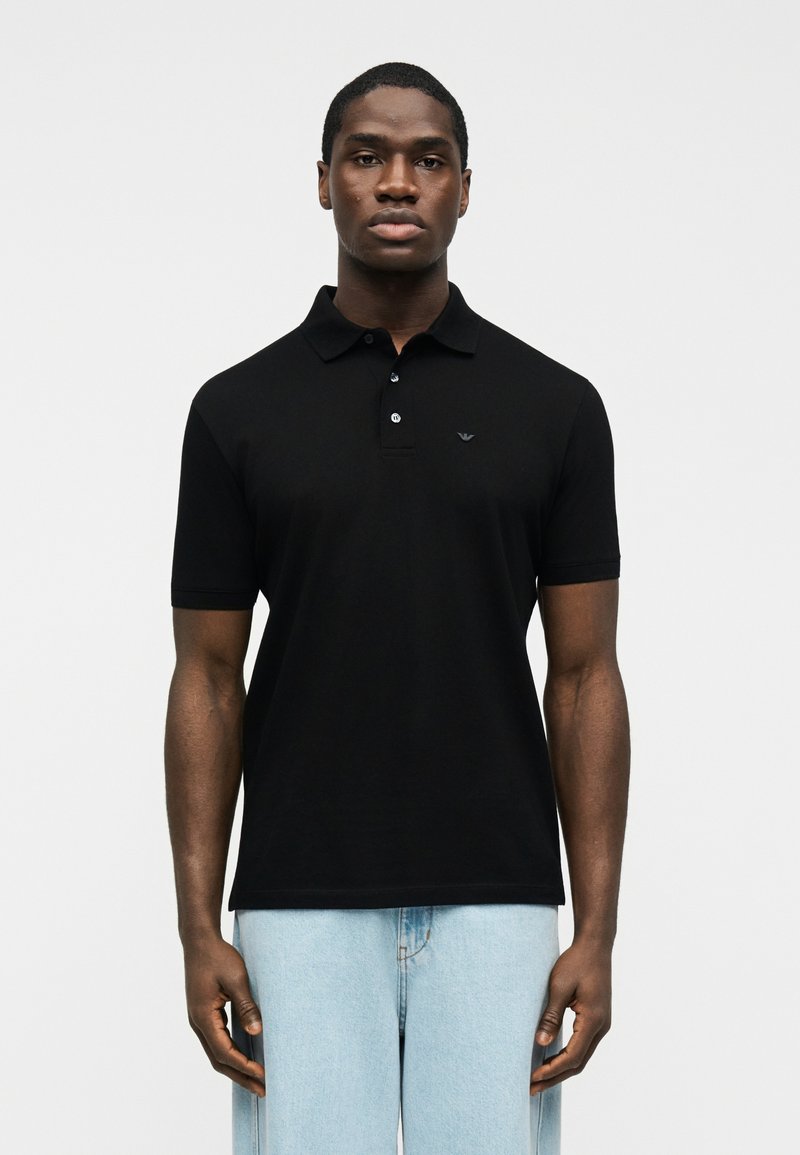 Άνδρας που φορεί μαύρο πόλο με κοντά μανίκια και ανοιχτό μπλε τζιν, στέκεται μπροστά σε απλό λευκό φόντο.