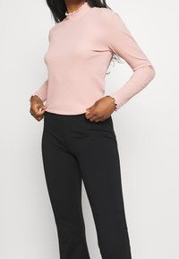 Woman wearing a light pink long-sleeve ribbed top with ruffled cuffs and high-waisted black pants, standing against a plain background.