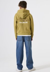 Persona con sudadera oliva con gráfico de montaña y el texto "TOTAL SOLITUDE" en la parte posterior, jeans azules y zapatillas blancas, de espaldas.