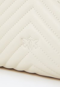 Beige ādas soma ar zīmuļu rakstu un kontrastējošām šuvēm. Iespējots uzspiestā logo detaļa apakšējā stūrī.