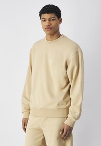 Sudadera beige con cuello redondo, mangas largas, puños acanalados y dobladillo. Presenta un pequeño logo bordado en la parte delantera. Textura suave y lisa.