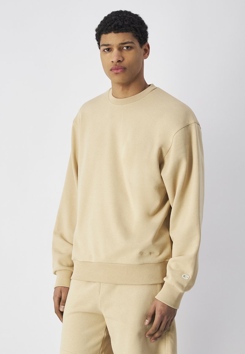 Sudadera beige con cuello redondo, mangas largas, puños acanalados y dobladillo. Presenta un pequeño logo bordado en la parte delantera. Textura suave y lisa.