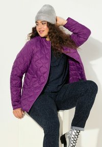 Giacca trapuntata viola con colletto alto, indossata sopra un top blu navy a trama. Pantaloni neri con motivo e stivaletti alla caviglia con lacci argentati. Cappellino grigio.
